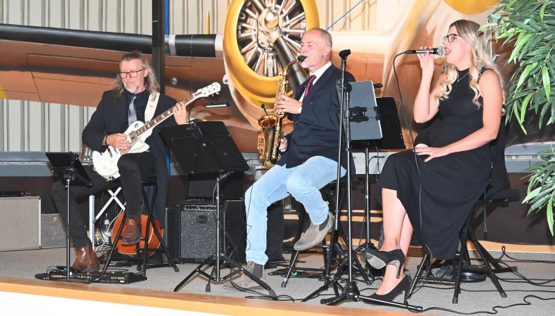Drei Musiker - Gitarrist, Saxophonist und Sängerin - sitzen musizierend auf der Bühne des Festsaals